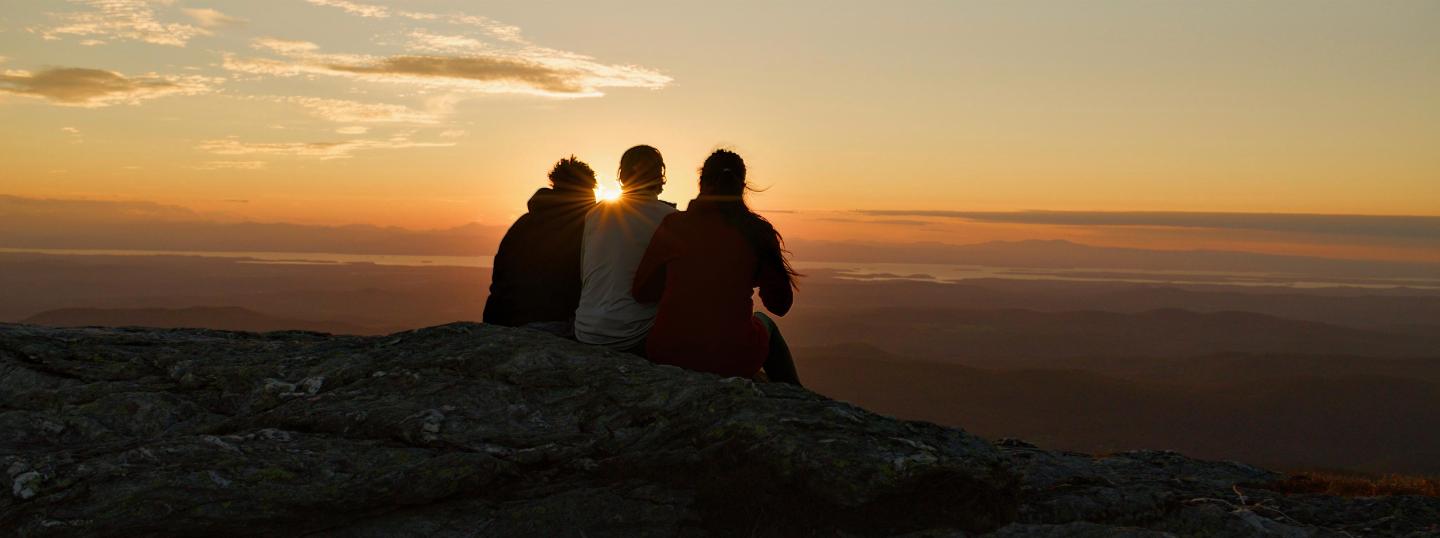 Sunset view with three people sitting on a rocky hilltop.