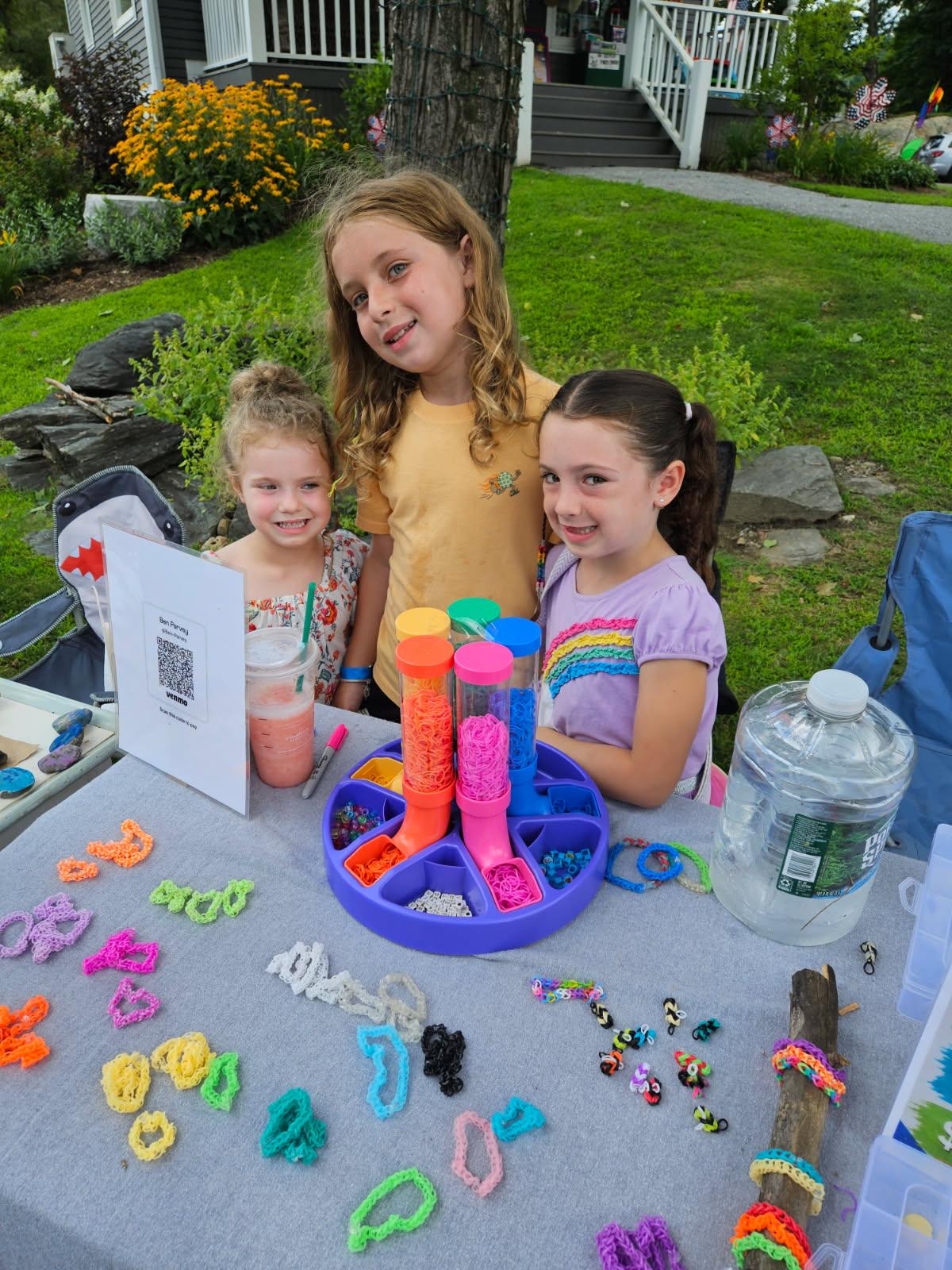Three children at a table with colorful crafts and a drink outside.