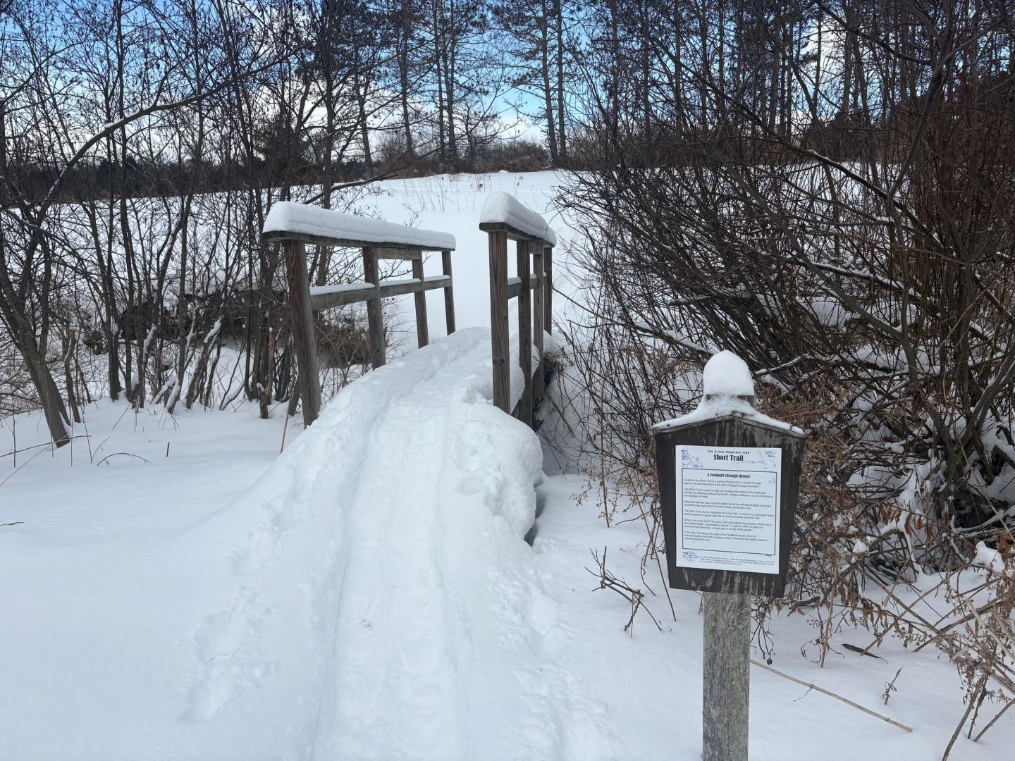 Snow-covered trail leading to a wooden bridge beside an informational sign in winter woods.