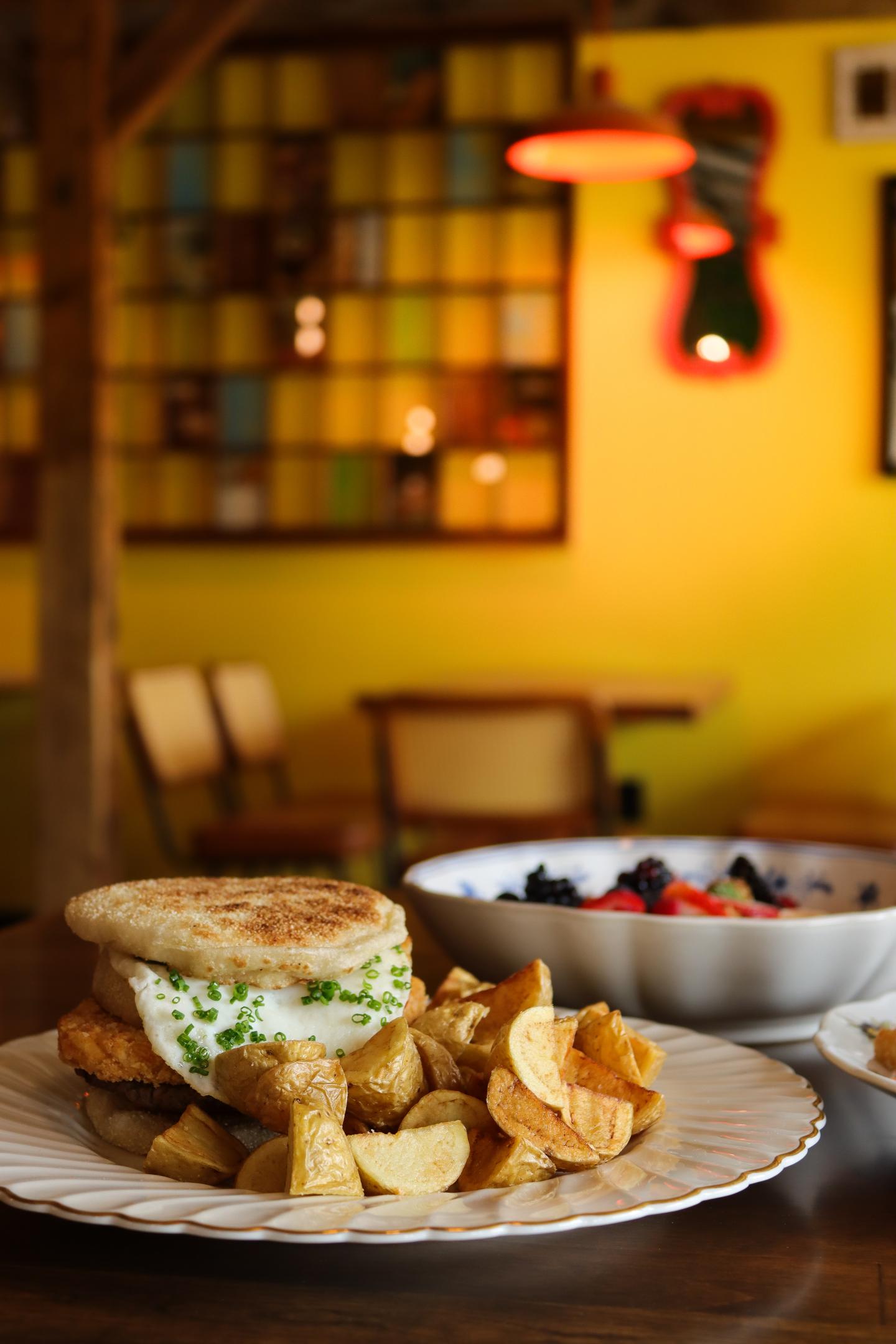 Breakfast sandwich with home fries and fruit bowl on a wooden table.