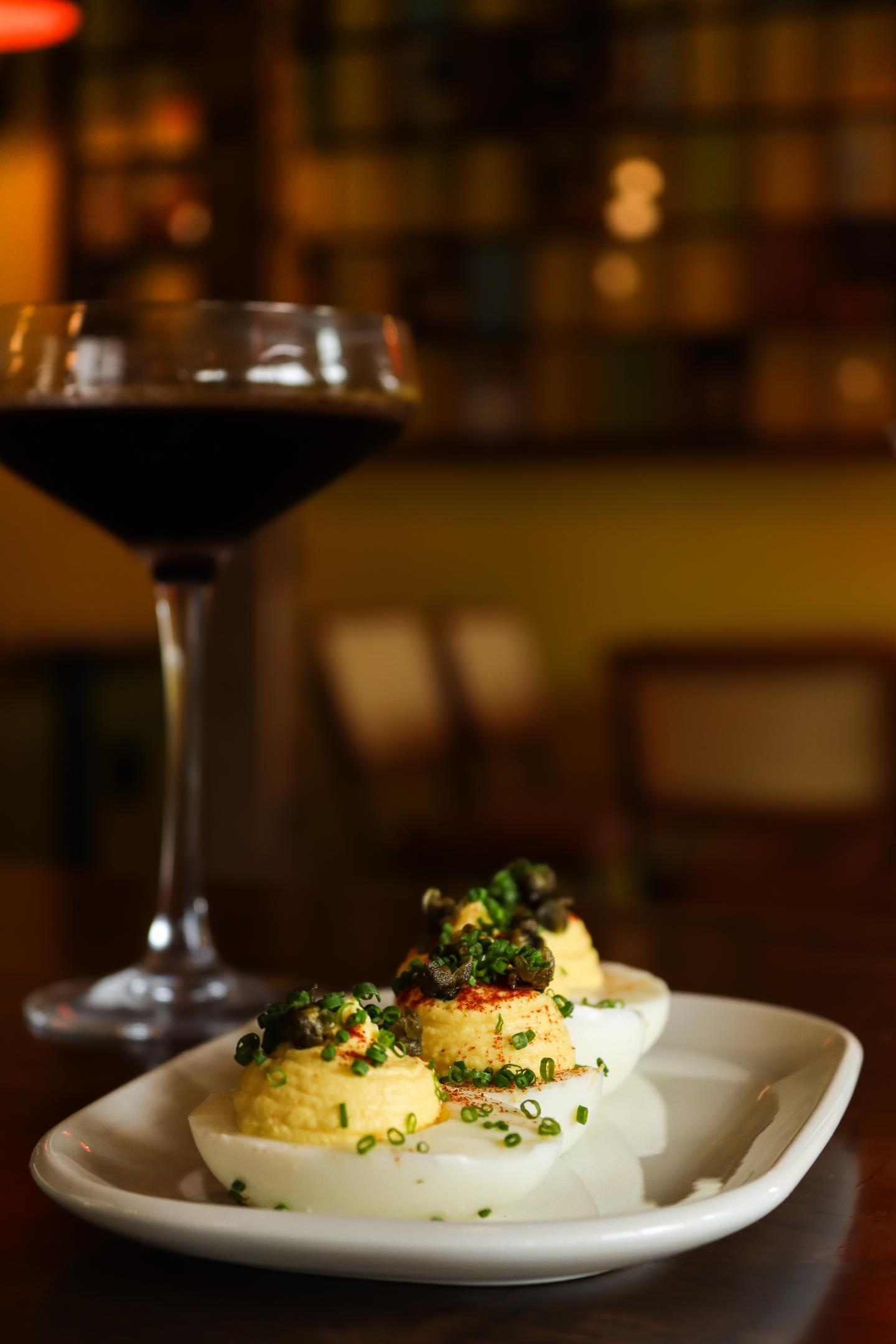 Deviled eggs with herbs on a plate, next to a glass of red wine in a dimly lit setting.
