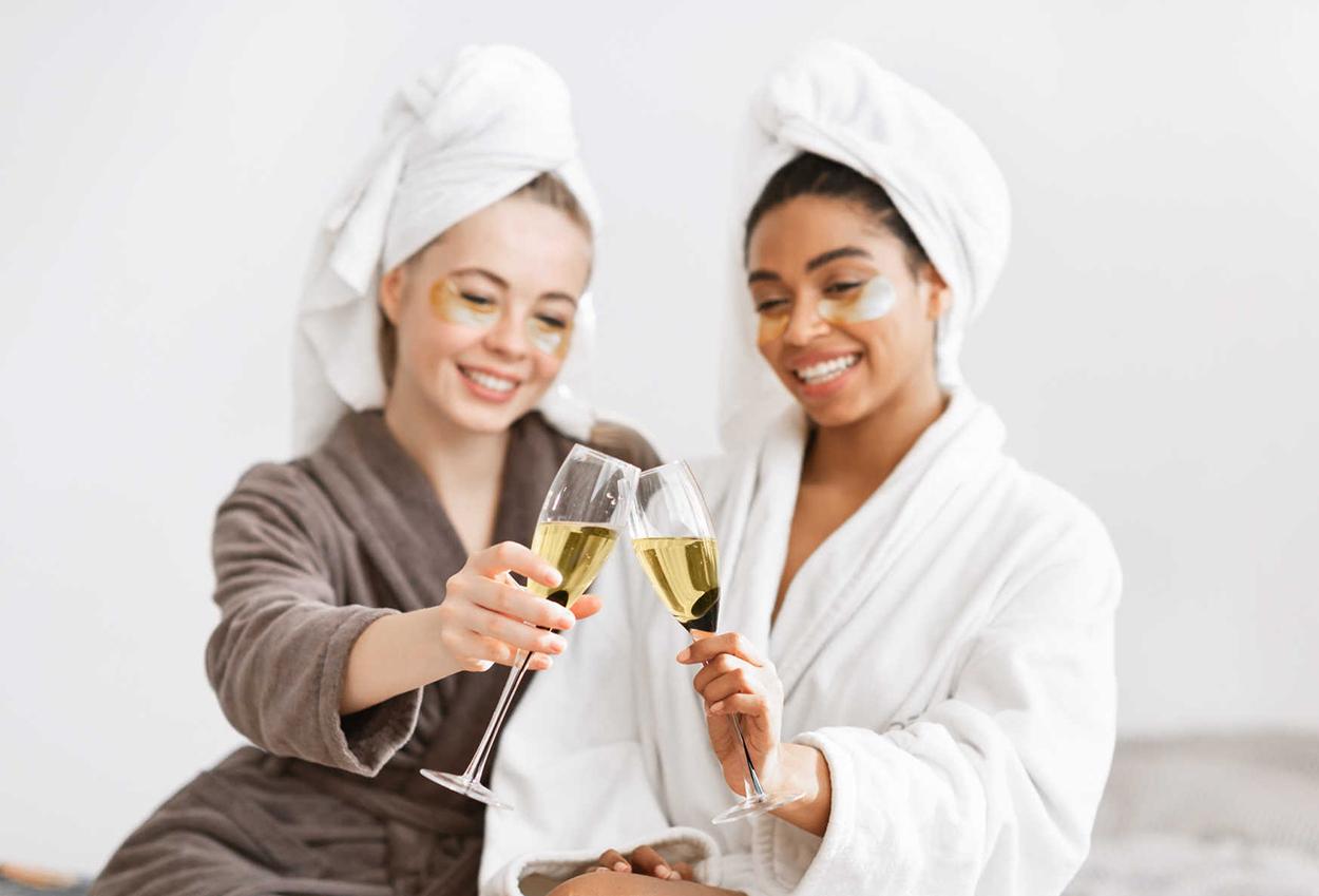 Two women in robes and towels toast with champagne glasses.