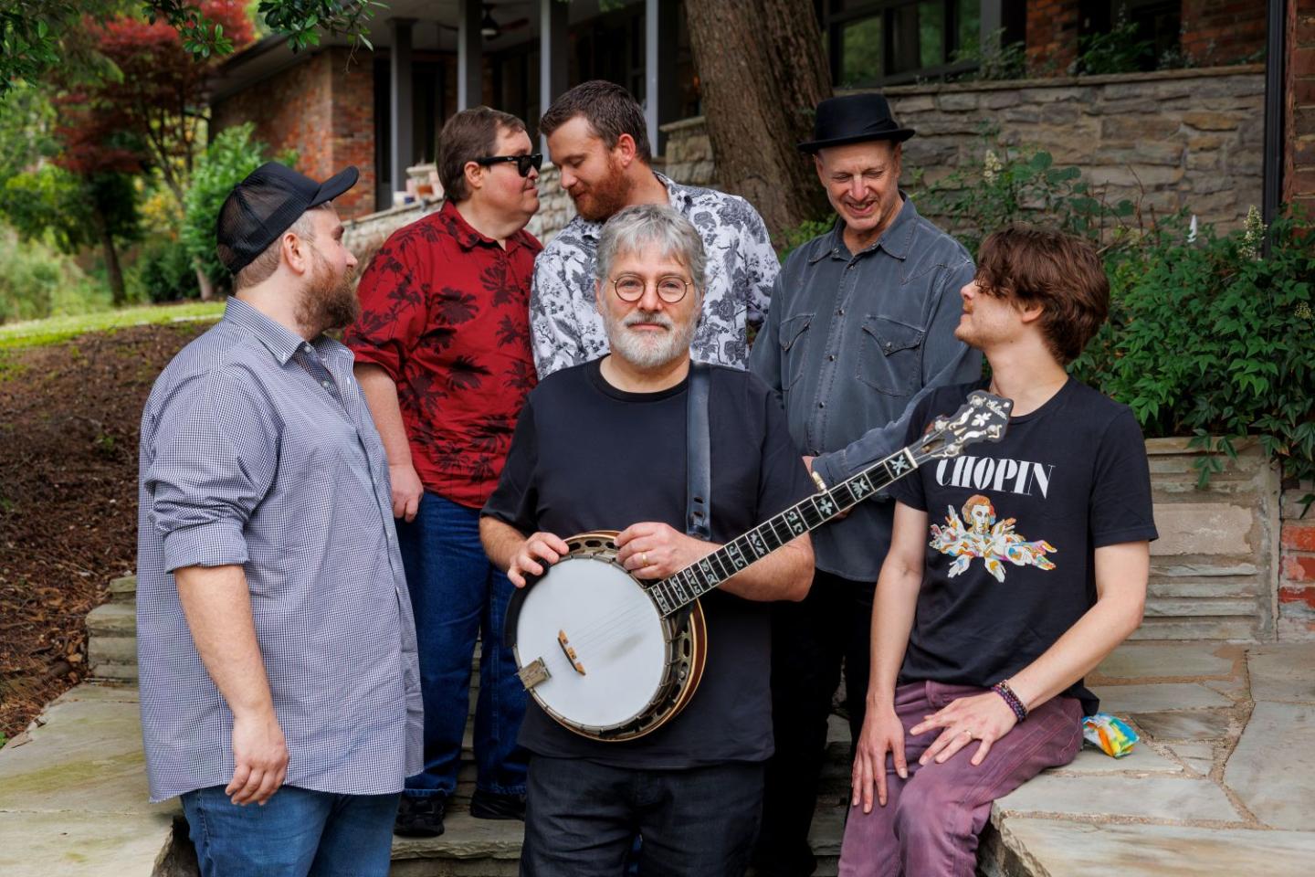 Five men outdoors, one playing a banjo while others watch.
