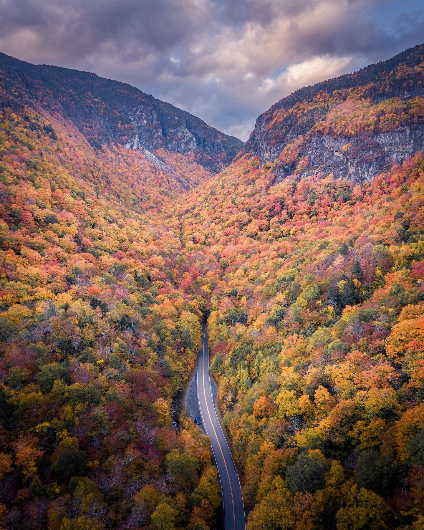 Winding road through a colorful autumn forest beneath mountains and cloudy skies