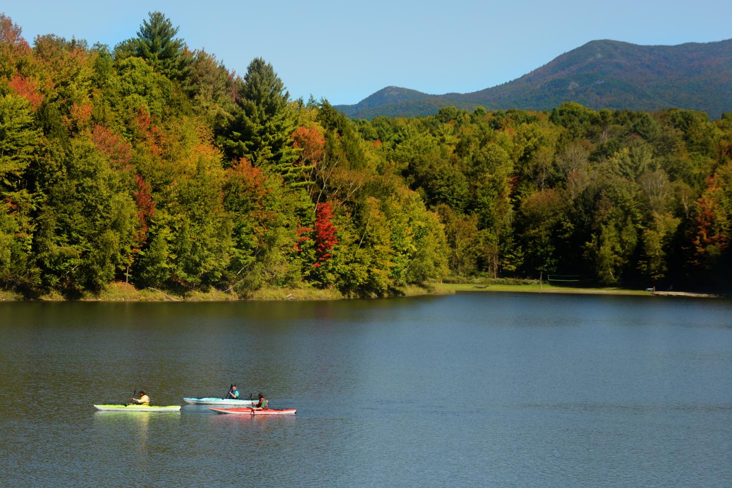 Three kayakers on a calm lake beneath colorful autumn forested hills.