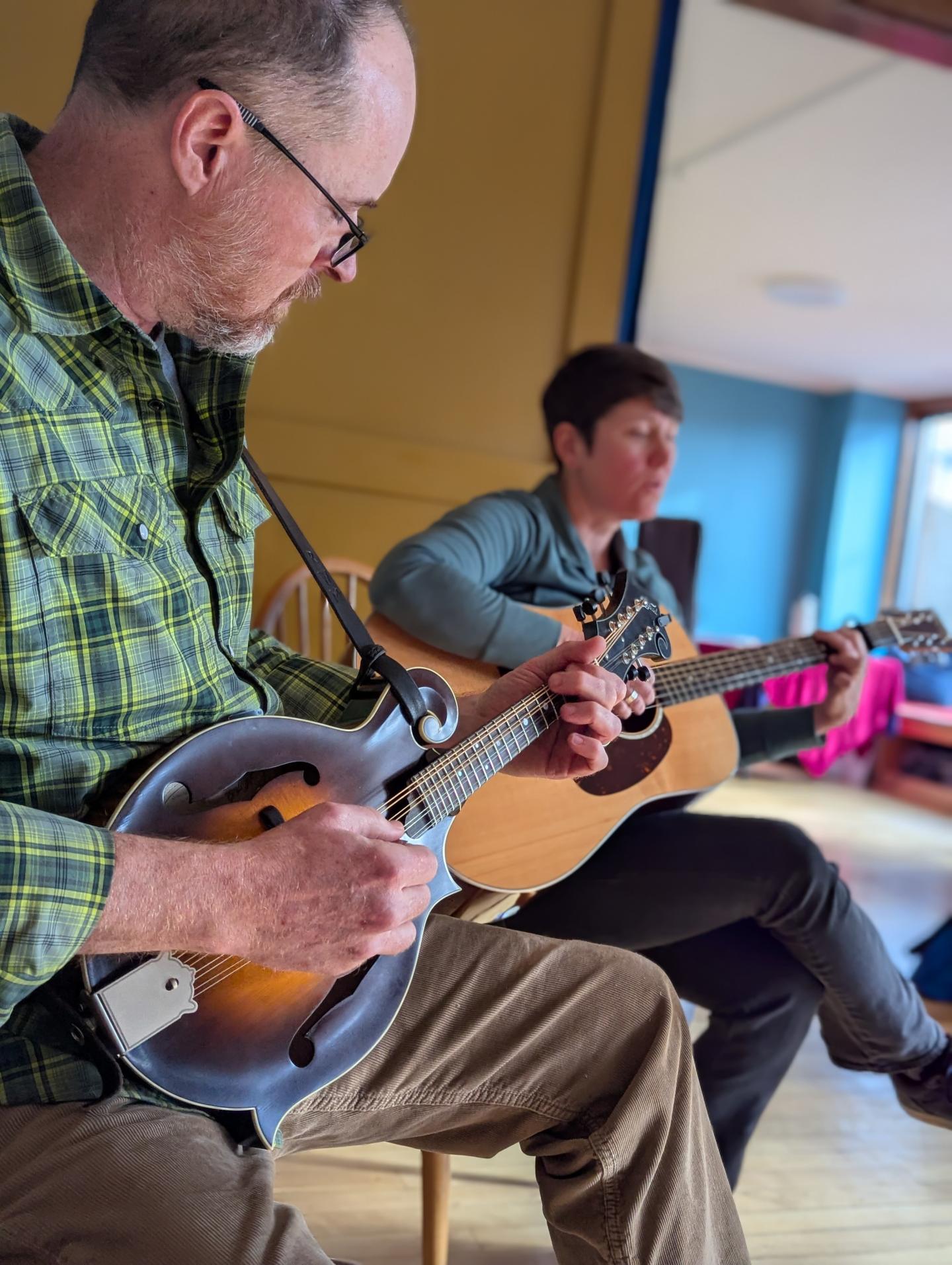 Two men playing string instruments indoors, one in green plaid and one in black.