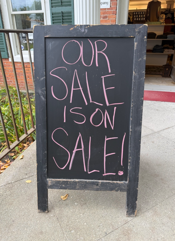 Black sidewalk chalkboard sign reading “Our sale is on sale!” in pink chalk.