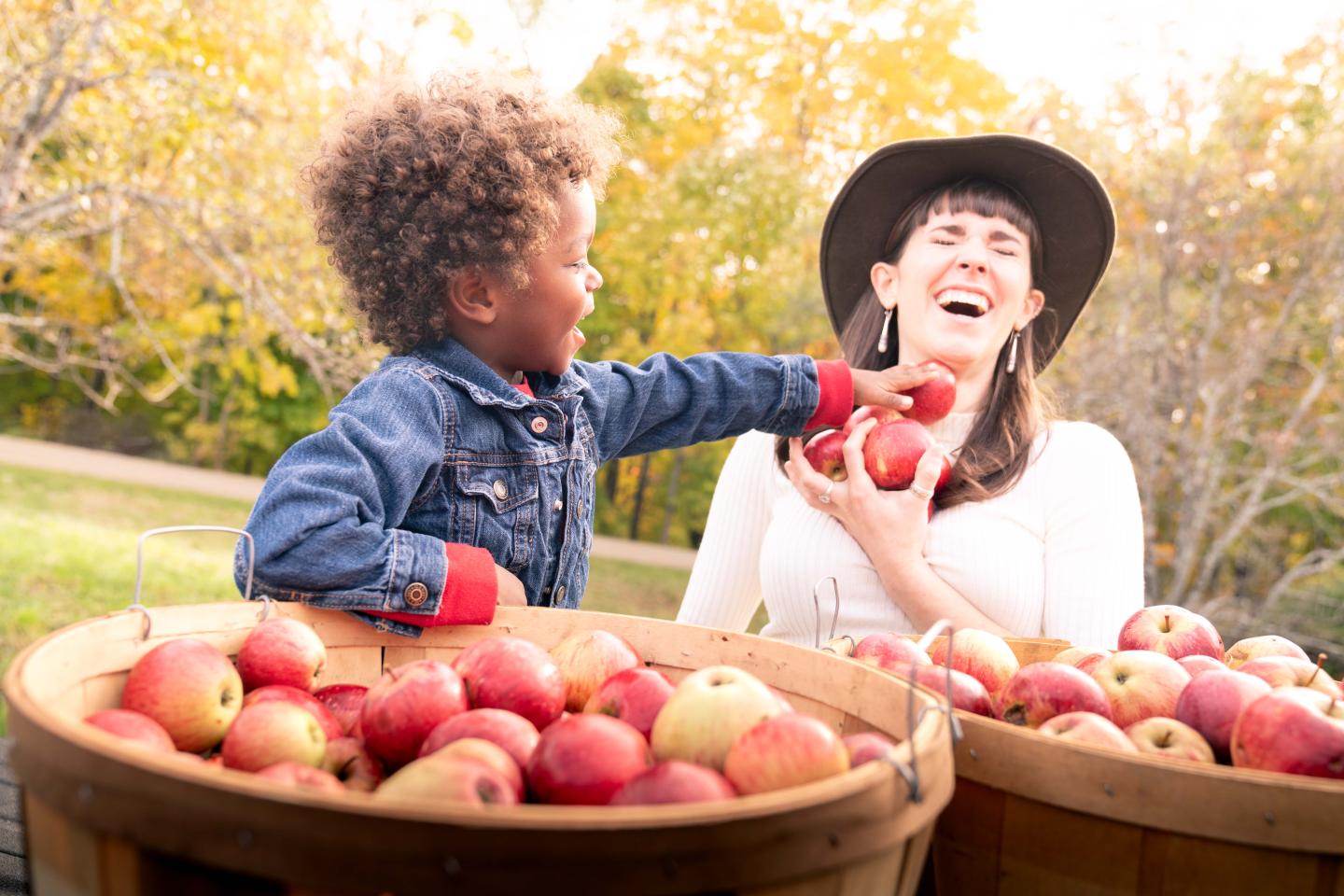 Child feeds an apple to a smiling woman beside baskets of apples.