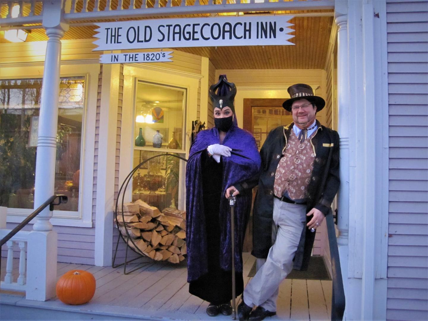 Two people in Victorian costumes stand outside the Old Stagecoach Inn at dusk.