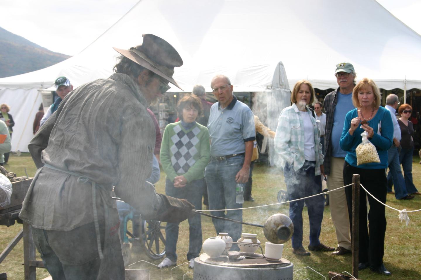 Blacksmith in historical clothing works at a smoky forge as visitors watch.