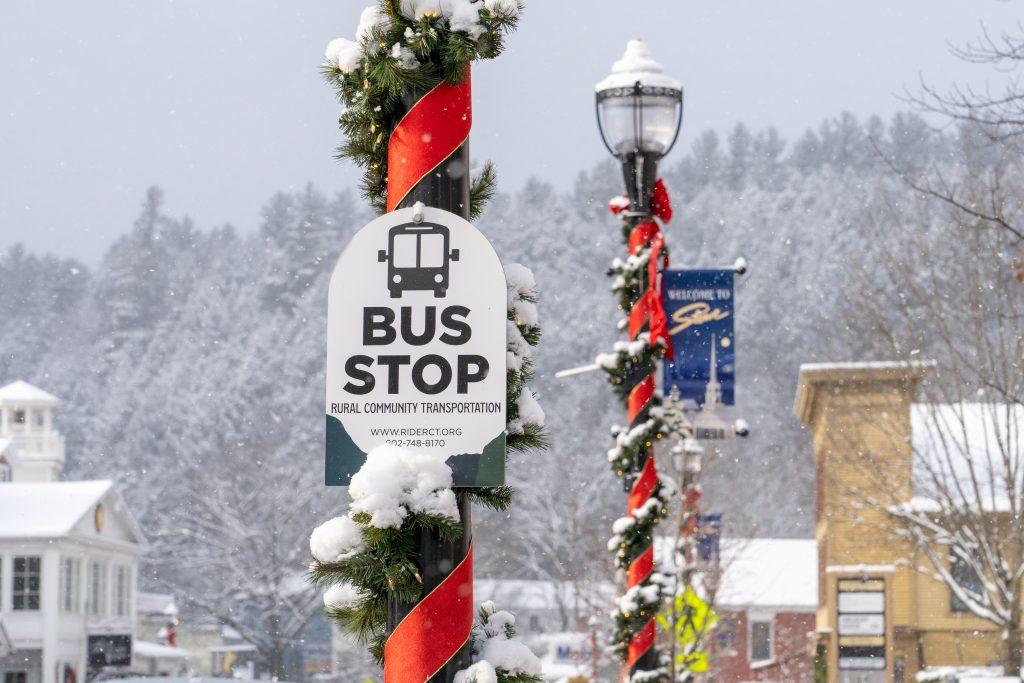 Snow-covered bus stop sign decorated with red ribbon and Christmas garland.