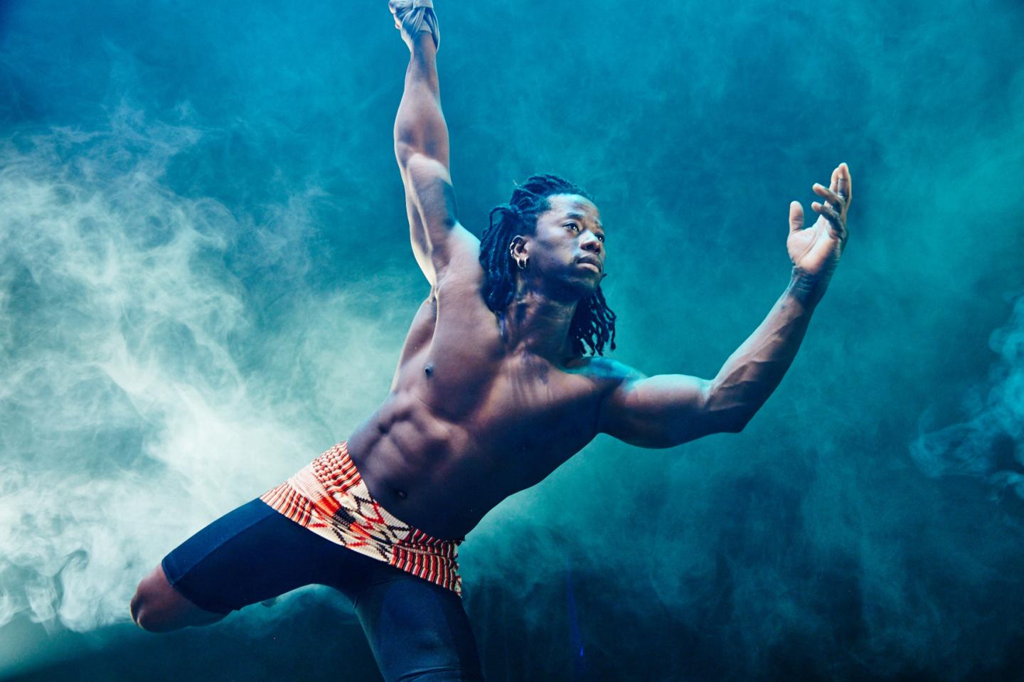 Shirtless male dancer leaping dramatically through blue and white smoke
