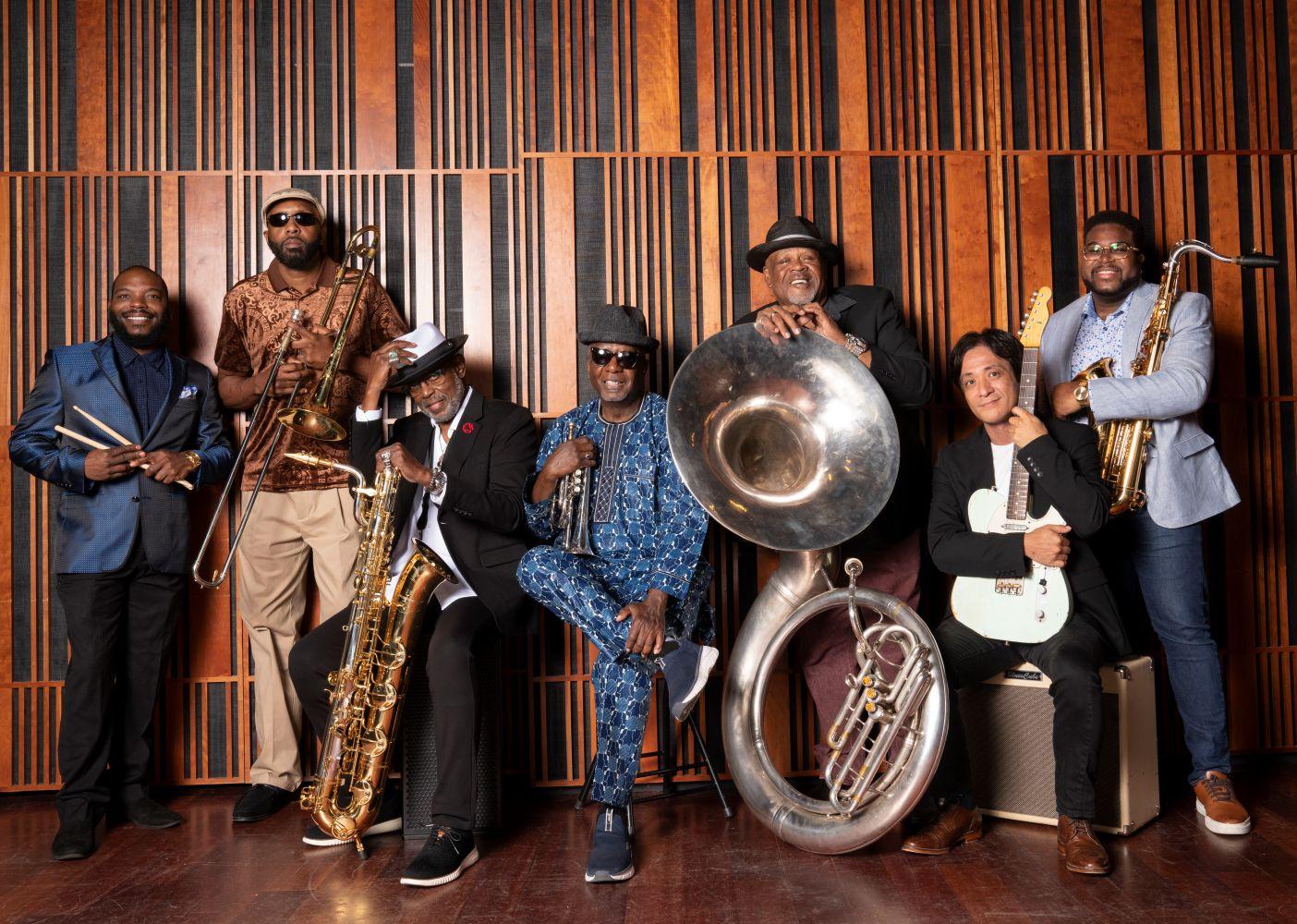 Seven musicians in formal attire pose with brass instruments against a wood-paneled wall.