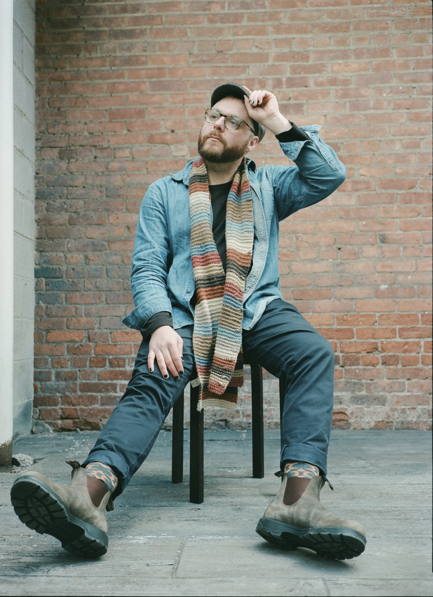 Man in denim and scarf sitting on a chair against a brick wall.