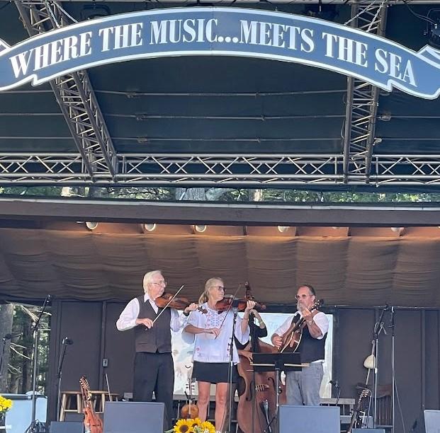 Three musicians perform on an outdoor stage beneath a “Where the Music Meets the Sea” banner.