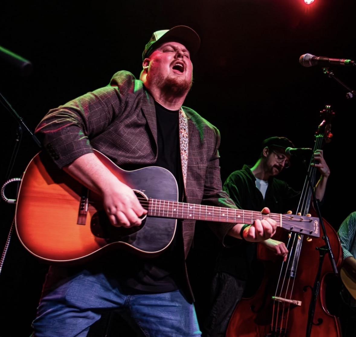 Bearded musician sings while playing acoustic guitar onstage.